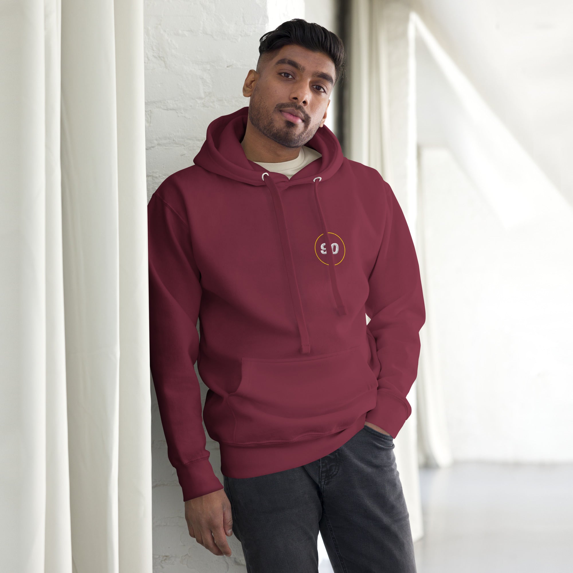 Man wearing a maroon hoodie with a logo, standing against a white wall | colour:maroon